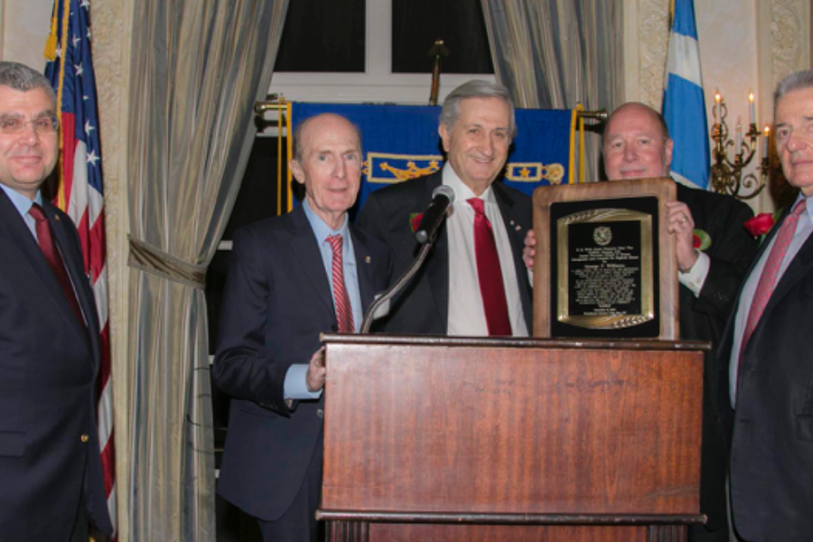 Five men in suits stand at a wooden podium at a formal event, one holding an engraved plaque, with an American flag and draped curtains behind them.