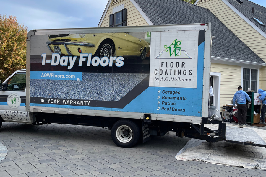 Box truck parked in a driveway with lettering reading "1-Day Floors," "Floor Coatings by A.G. Williams," and "15-Year Warranty," as two workers unload materials near a house.