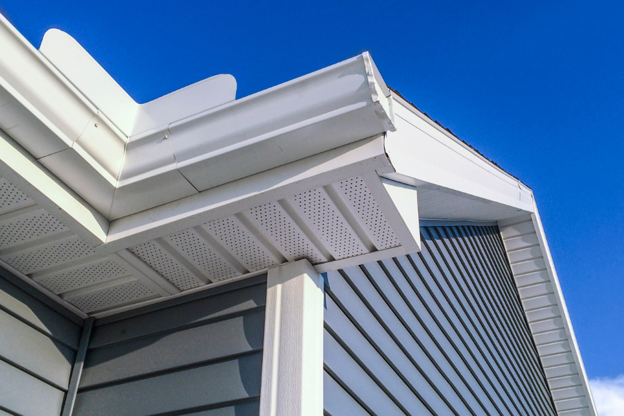 Corner of a house with light gray siding, white PVC trim, gutters, and vented soffit panels against a bright blue sky.