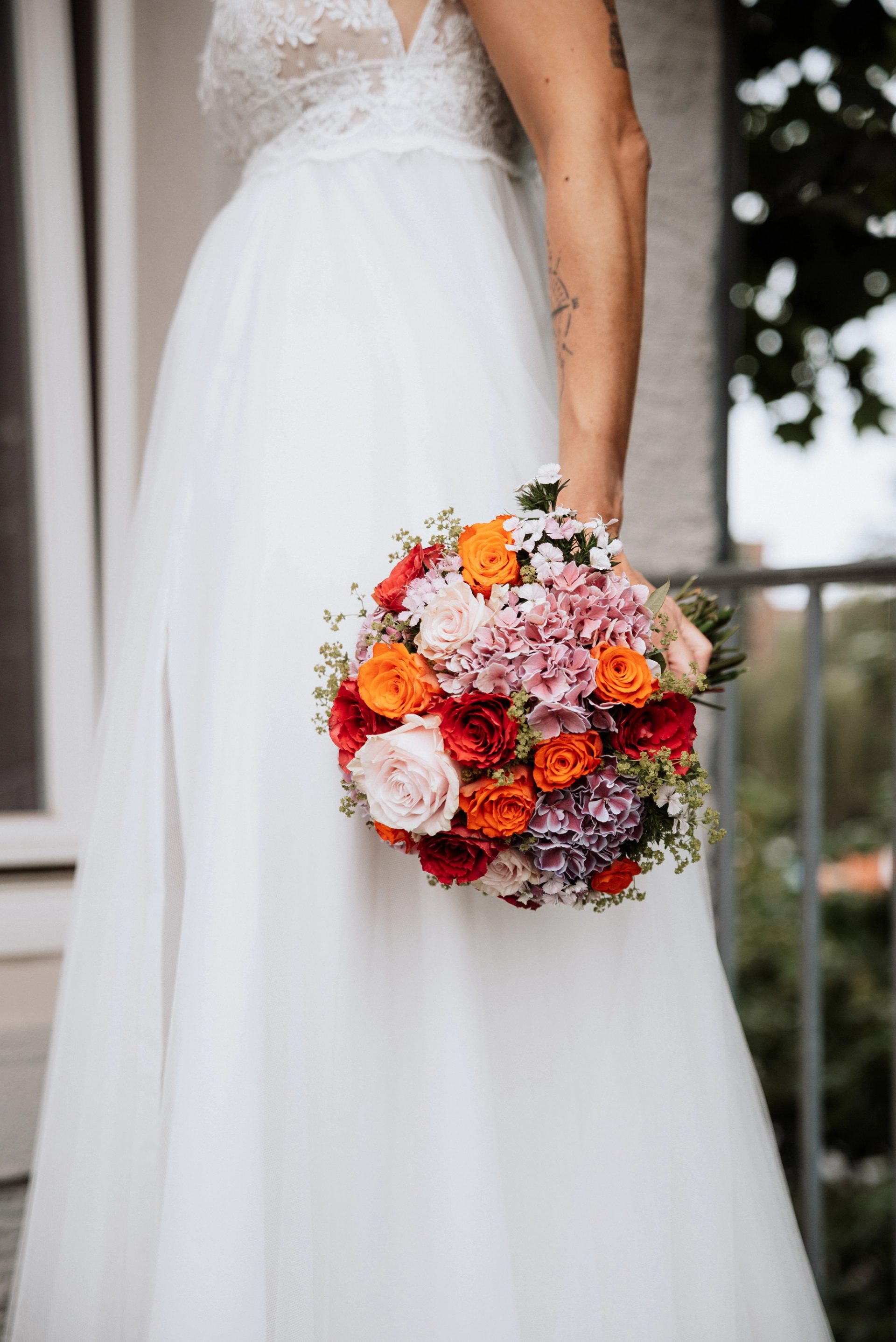 Der bunte Brautstrauß aus Rosen und Hortensien in Marens Hand vor dem weißen Tüllrock