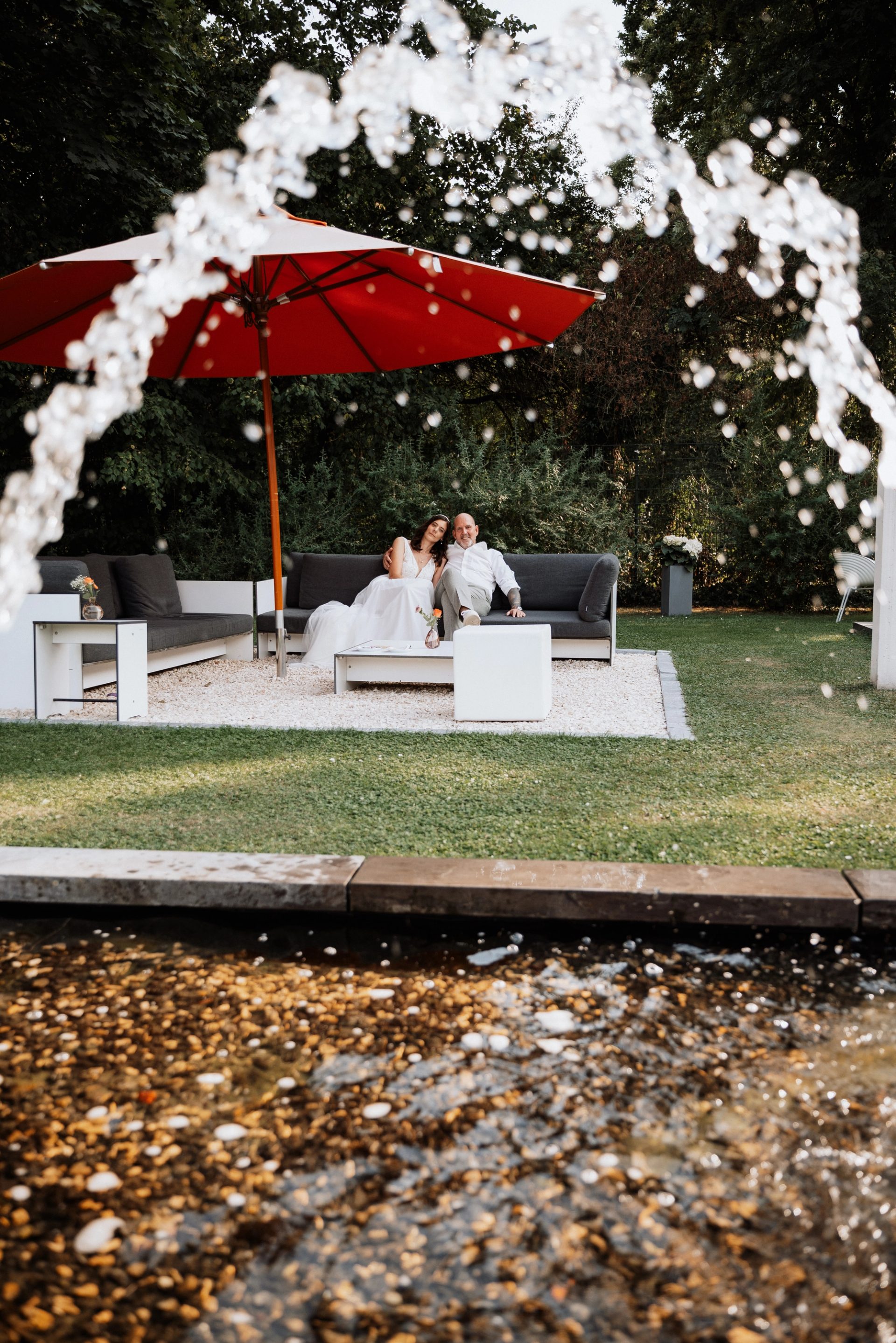 Maren und Ralph auf der Gartenlounge, davor Wasserstrahlen vor einem roten Sonnenschirm