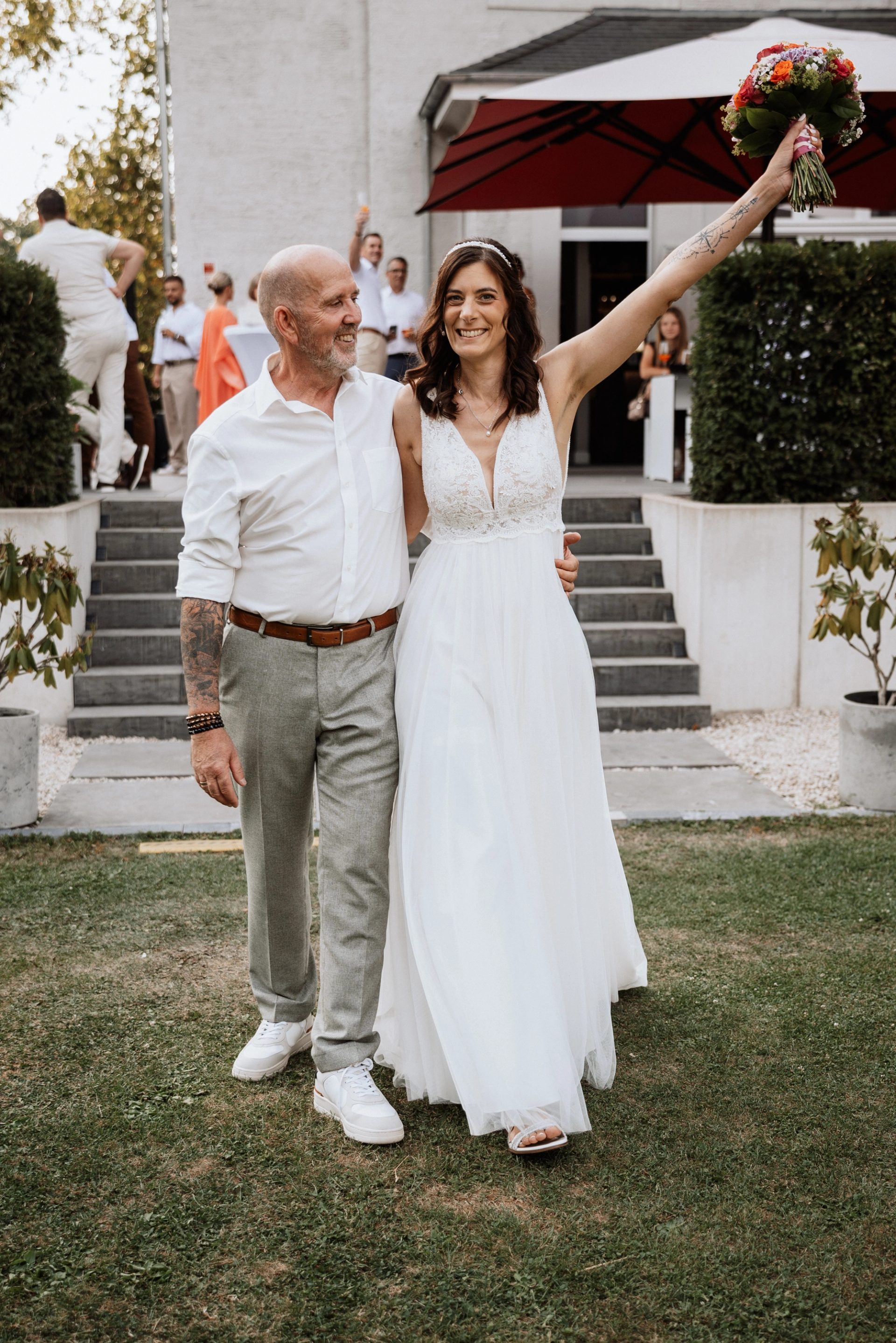 Maren reckt lachend den Brautstrauß in die Höhe, Ralph schaut sie an, hinter ihnen die Terrasse der Villa Glanzstoff