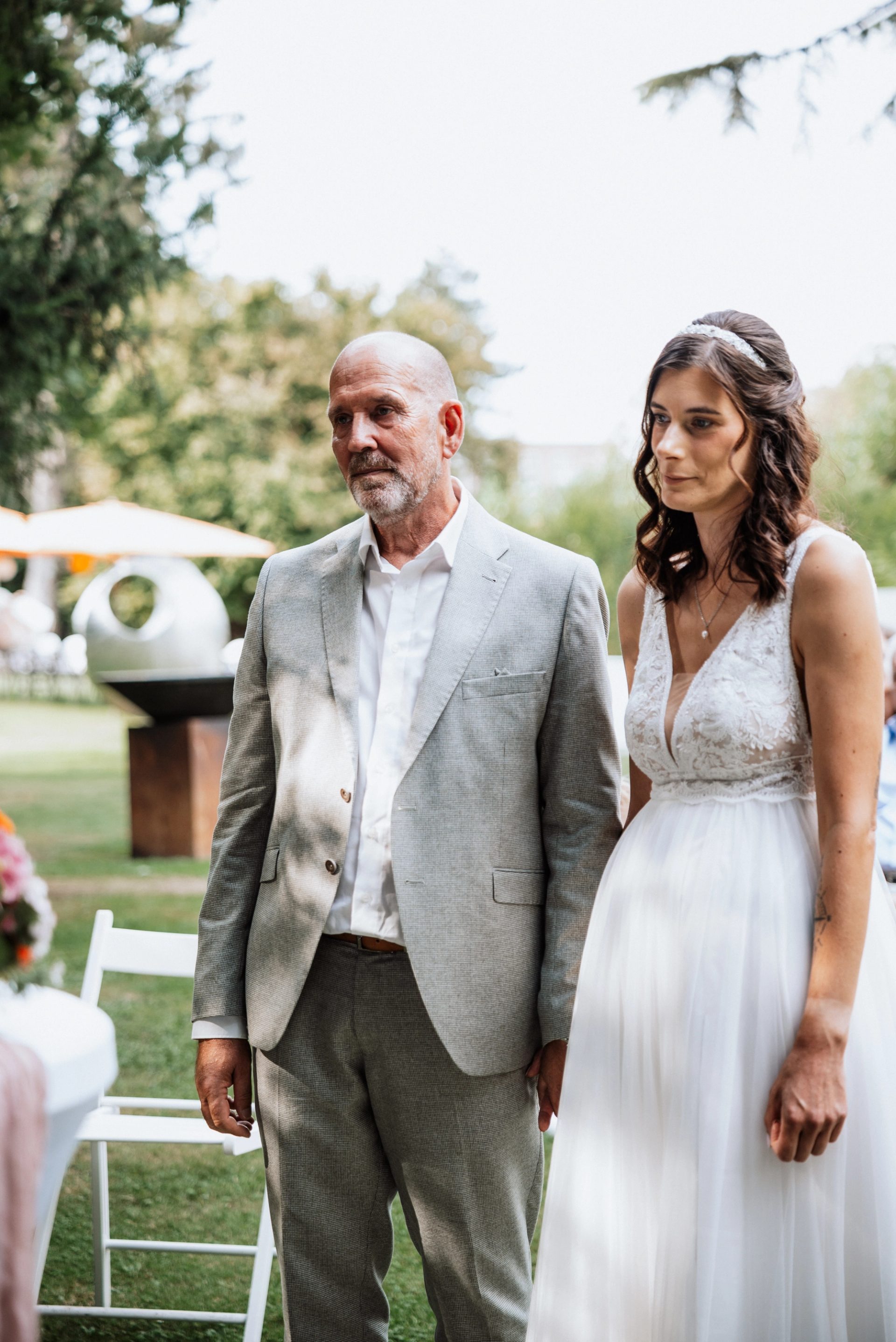 Ralph und Maren stehen nebeneinander im Garten der Villa Glanzstoff, neben ihnen weiße Stühle