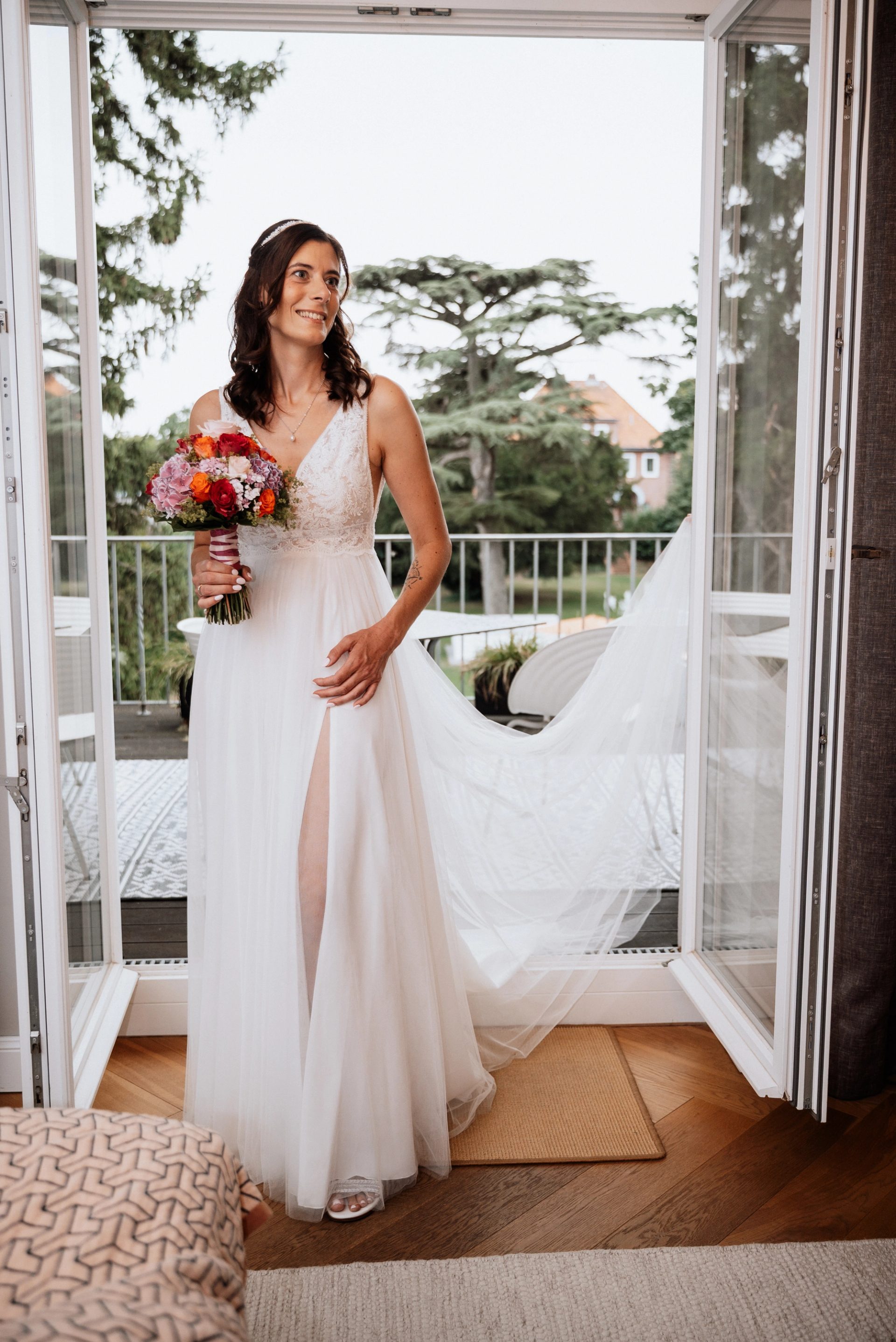 Maren im Brautkleid lächelt in der offenen Balkontür der Villa Glanzstoff, den Strauß in der Hand