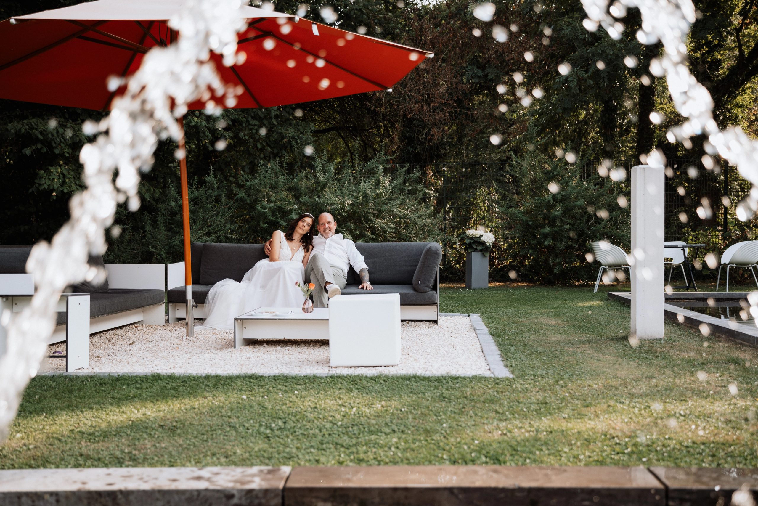 Die Gartenlounge der Villa Glanzstoff mit rotem Sonnenschirm, Maren und Ralph sitzen eng beieinander, im Vordergrund Wasserstrahlen