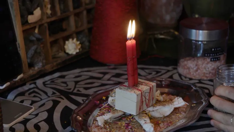 A red candle burning on a box wrapped in red thread, on a plate of herbs and petals