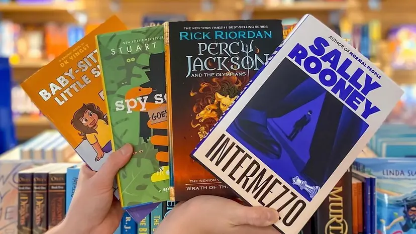 Two hands hold up four young adult books. There are shelves of more books in the background.