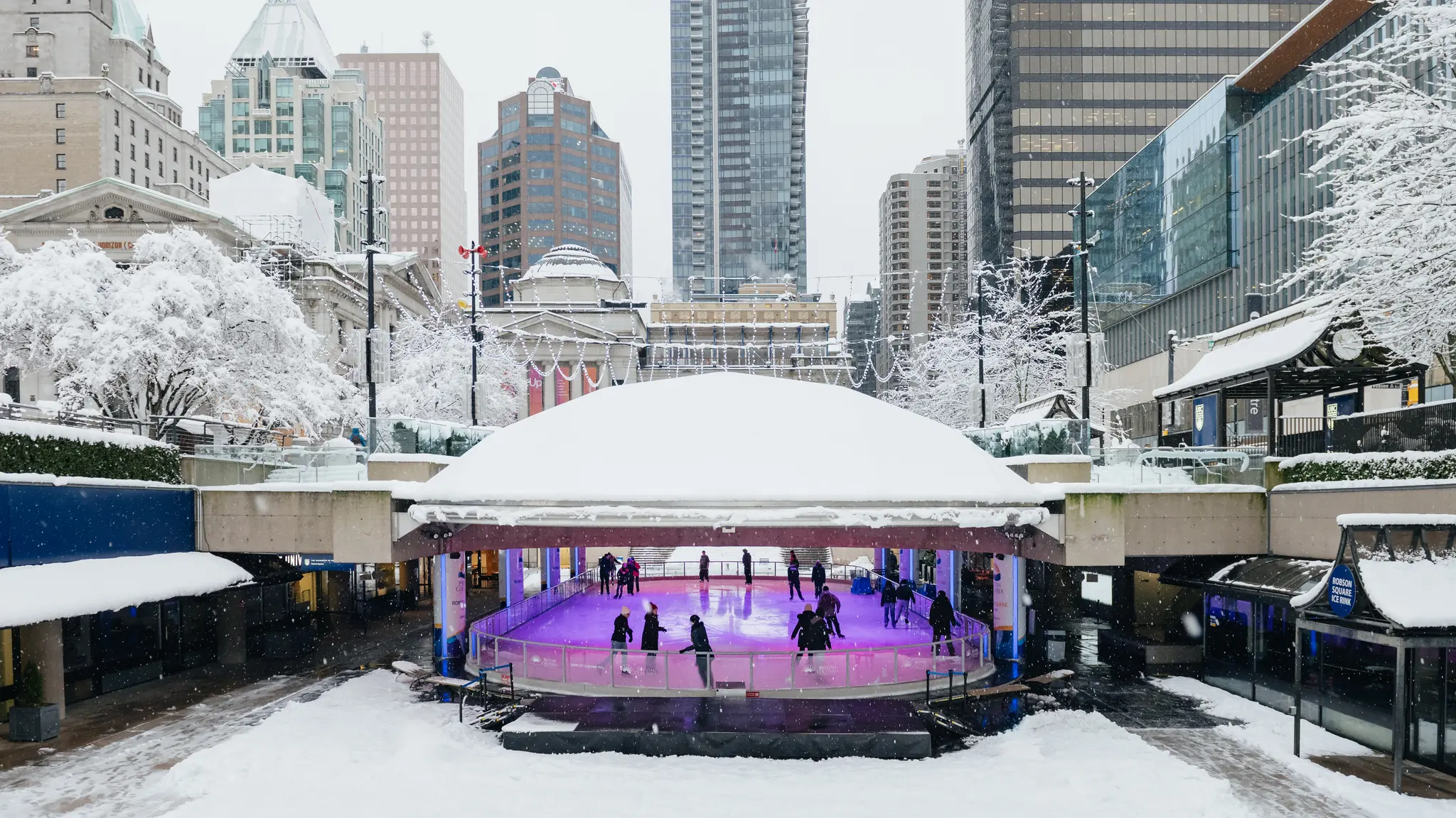 Skaters under the dome at the Robson Square Ice Rink. The surrounding city is covered in snow.