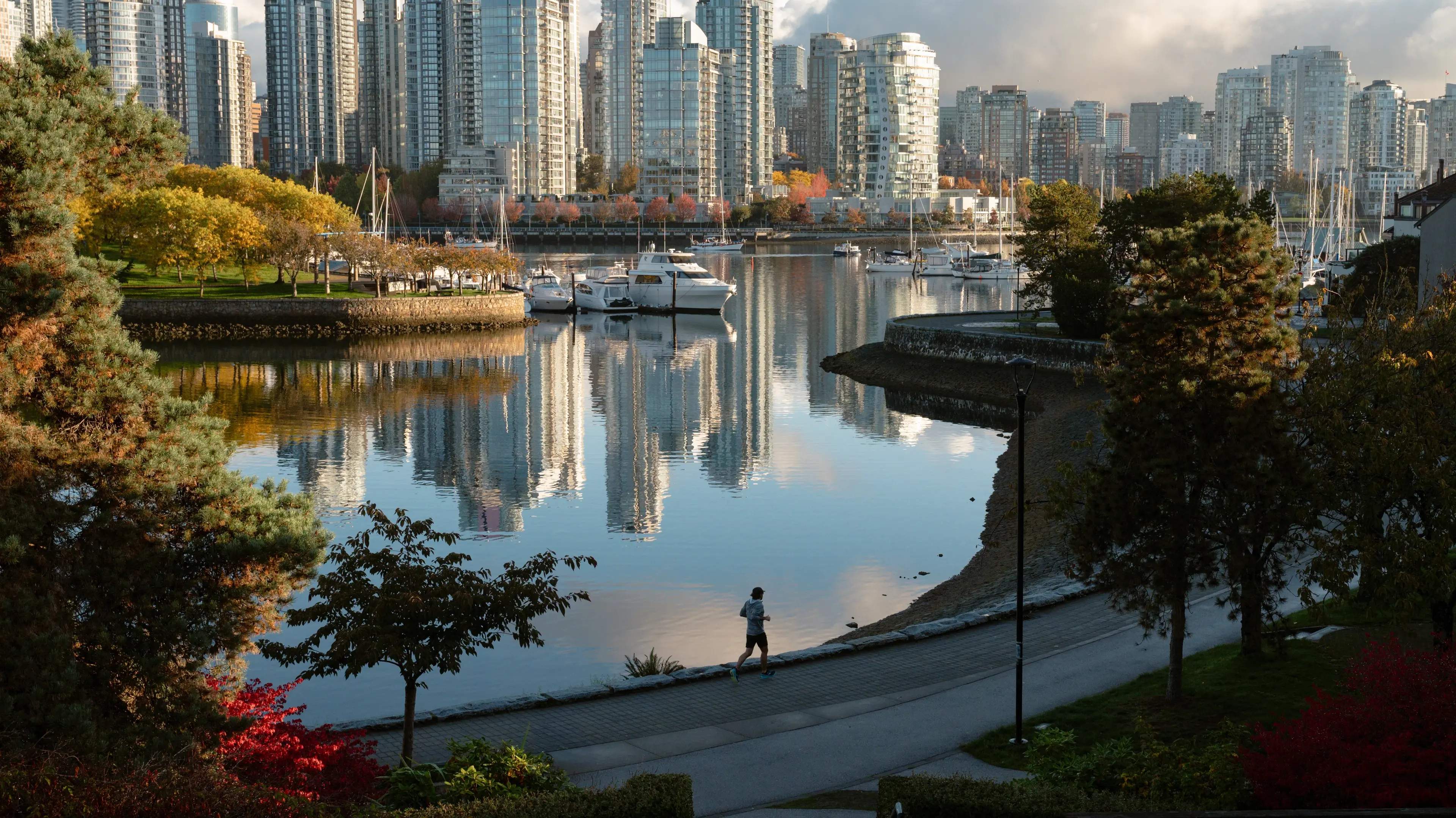 Person running along the seawall in False Creek with the downtown Vancouver skyline in the background, framed by trees in vibrant fall colours beside the water.