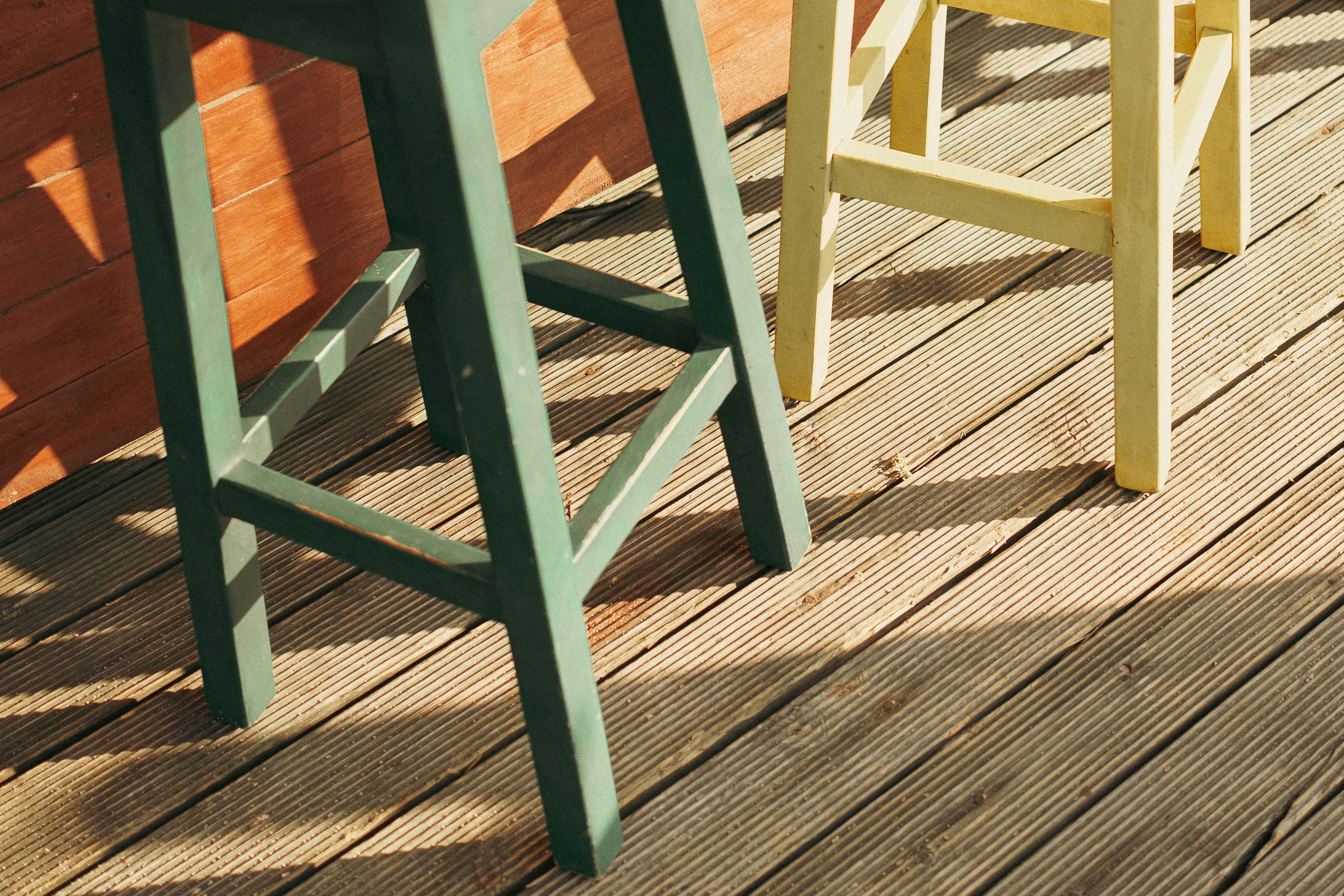 Green and yellow painted wooden bar stools on a weathered wood deck in North Jersey, showing sun-faded decking boards in need of repair or replacement.