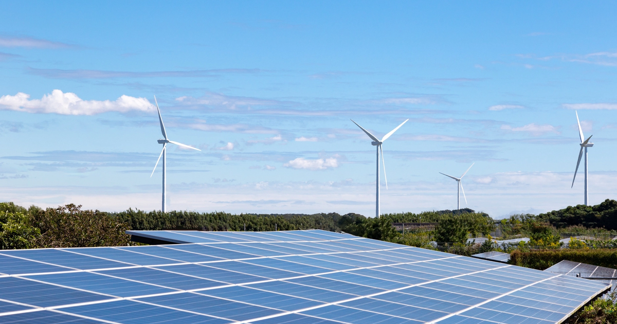 Solar panels in the foreground, with wind turbines in the background, generating renewable energy under a clear blue sky, suggesting they reach businesses through energy brokers and direct suppliers.