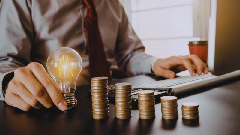Person working at a desk with rising stacks of coins and a light bulb to emphasise fixed vs variable electricity rates.