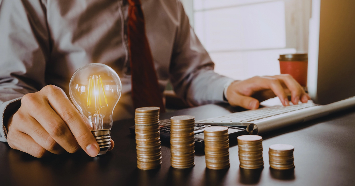 Person working at a desk with rising stacks of coins and a light bulb to emphasise fixed vs variable electricity rates.