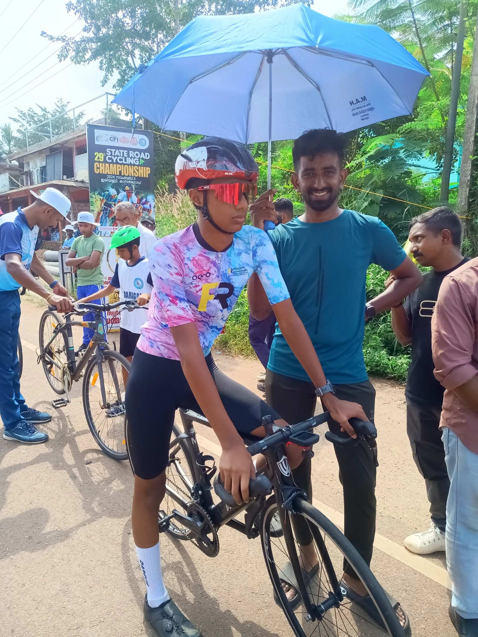 Juan Joy with fellow cyclist Saiju DS at a road cycling event, standing beside Juan's bicycle under a blue umbrella.