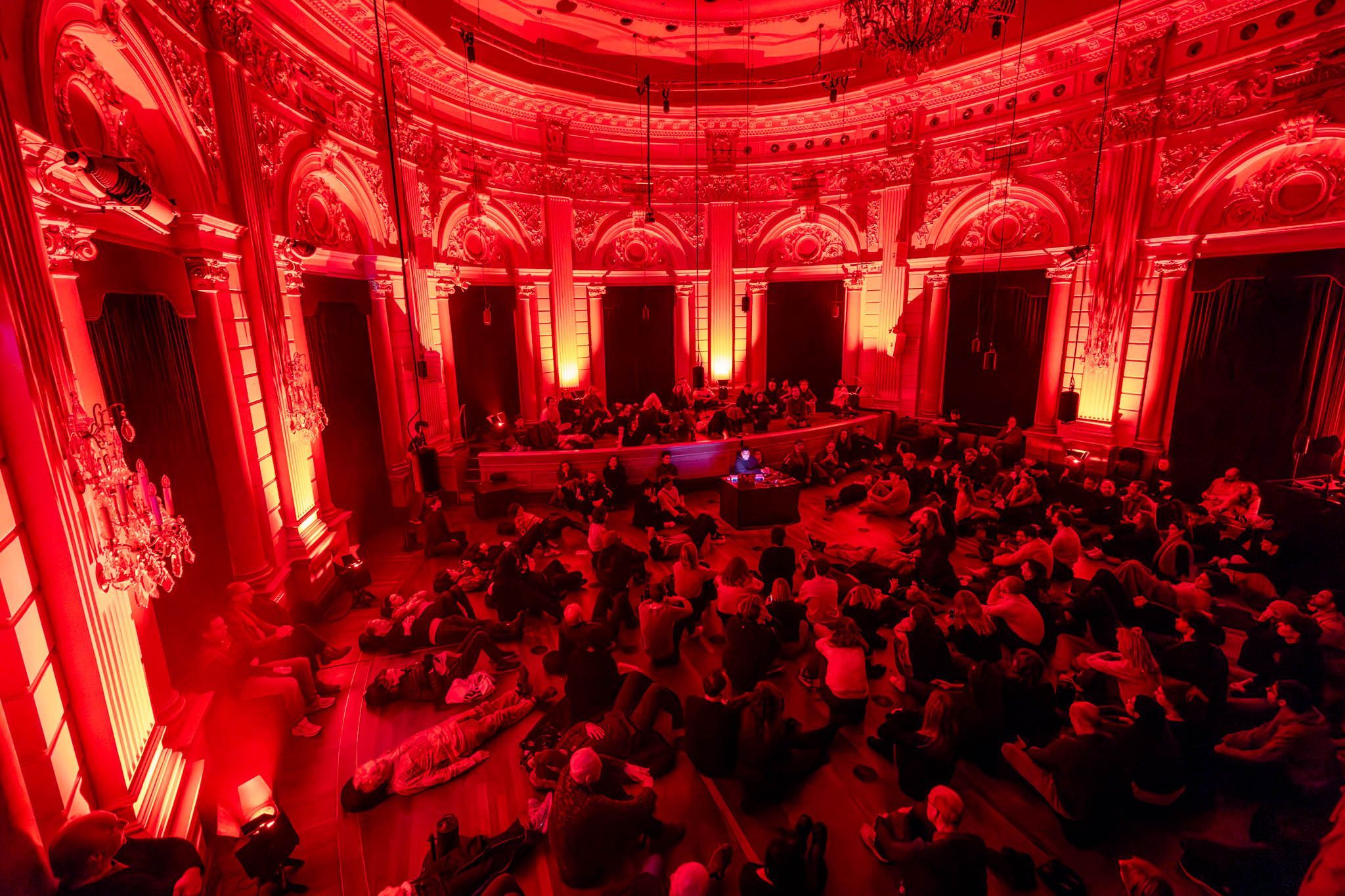 A group of people sitting and lying, listening together, in a dramatically lit room with a strong red hue. 