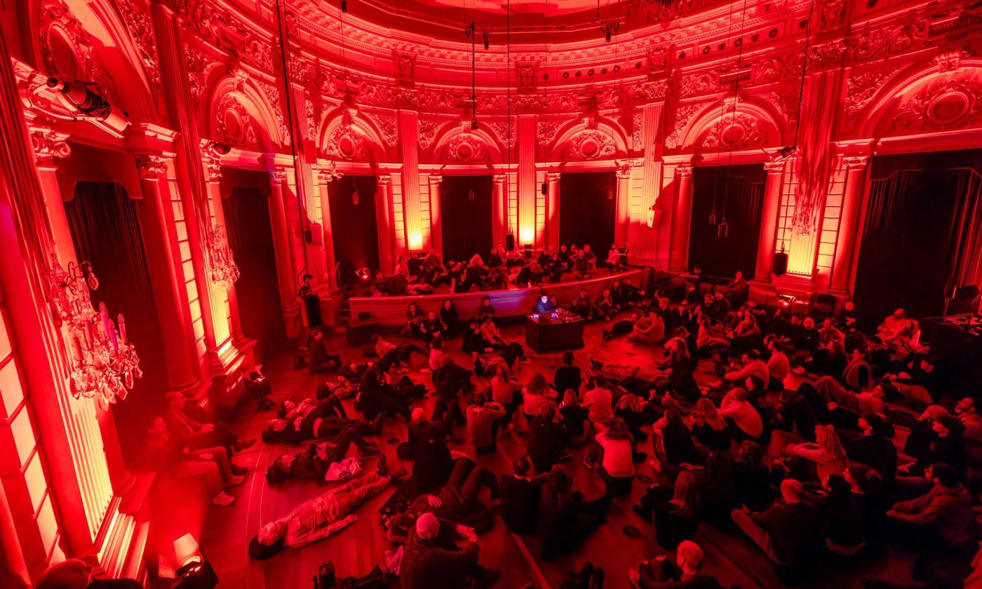A group of people sitting and lying, listening together, in a dramatically lit room with a strong red hue.