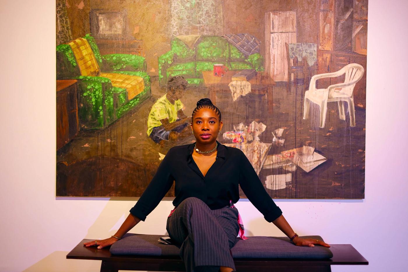 Alt= “the curator Jennifer Agusiobo is wearing a black shirt and grey trousers. She has jewelry on both wrists and her neck. She is seated in front of an art piece at the exhibition hall for The Rooms We Carry art show in Retro Africa Gallery, Asokoro, Abuja, Nigeria”