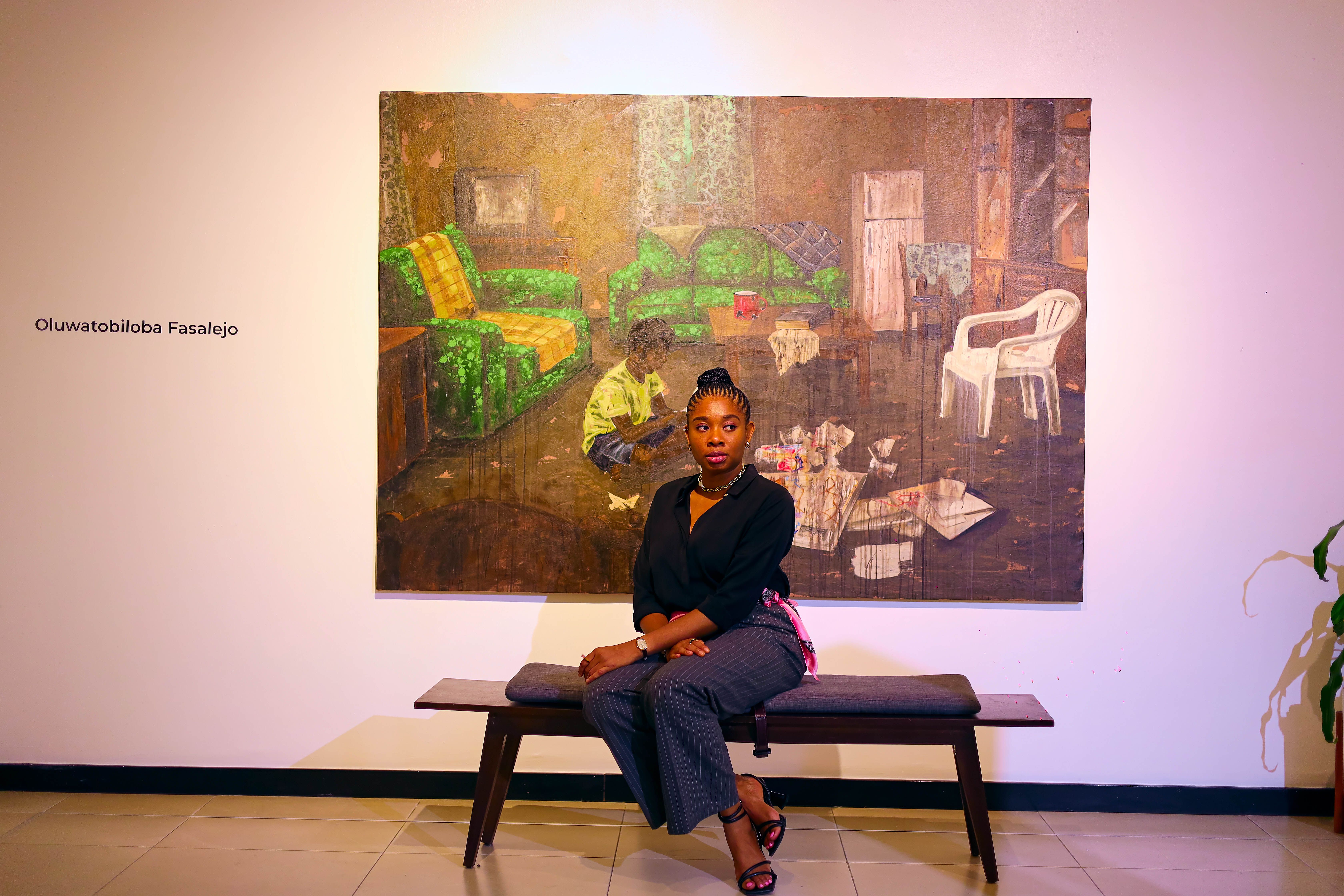 Th curator Jennifer Agusiobo is wearing a black shirt and grey trousers, she is seated in front of Oluwatobiloba Faselejo’s art work in Retro Africa Gallery