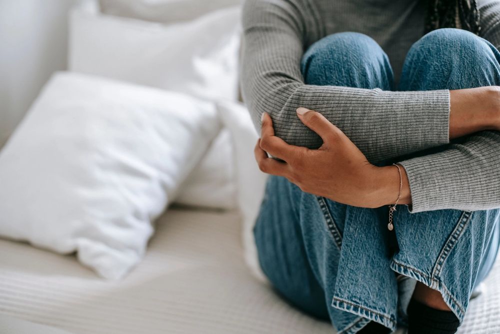 A person sits on a bed, hugging their knees to their chest, conveying sadness or distress.