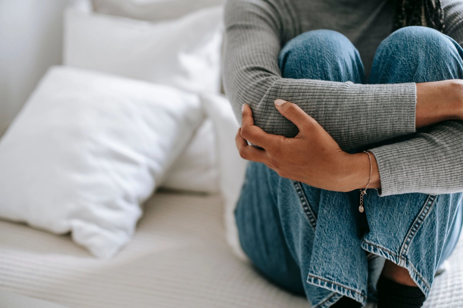 A person sits on a bed, hugging their knees to their chest, conveying sadness or distress.