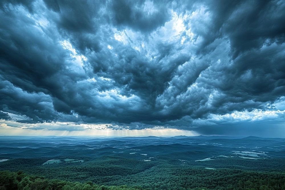 Dramatic dark storm clouds with sunbeams over a vast, green forested landscape.