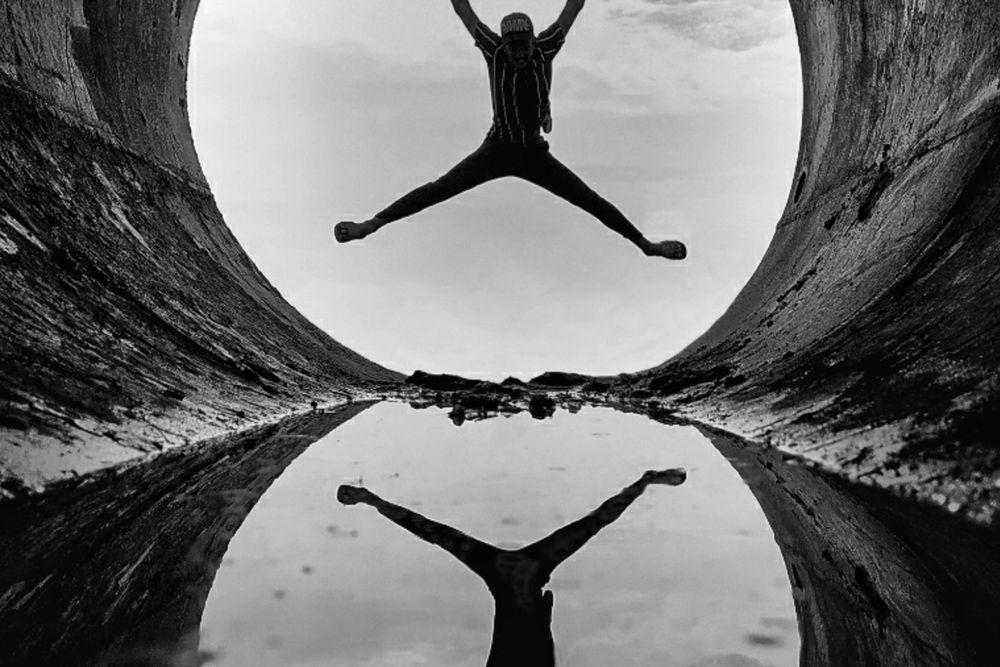 Black and white image of a person jumping with arms and legs spread, silhouetted against a cloudy sky within a circular opening, perfectly reflected in water below.
