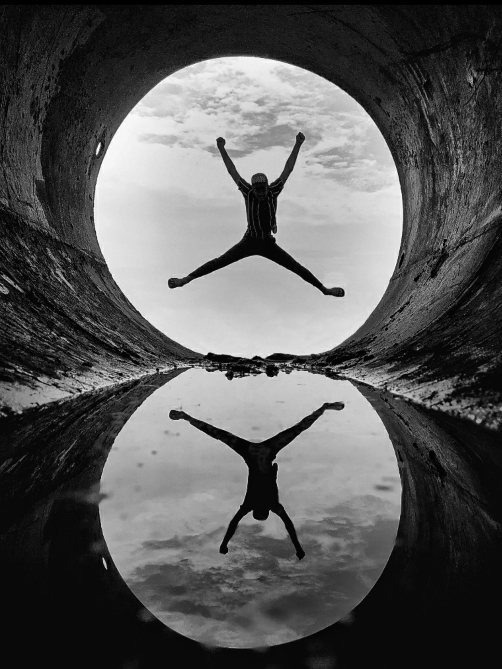 Black and white image of a person jumping with arms and legs spread, silhouetted against a cloudy sky within a circular opening, perfectly reflected in water below.