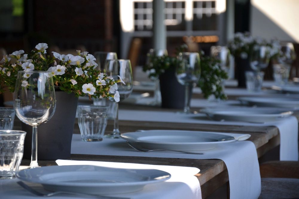 An outdoor dining table set with white plates, glasses, and potted white flowers on a wooden table.