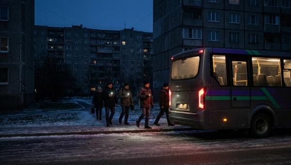 Працівники з ліхтариками телефонів ідуть до корпоративного автобуса біля панельних багатоповерхівок перед світанком