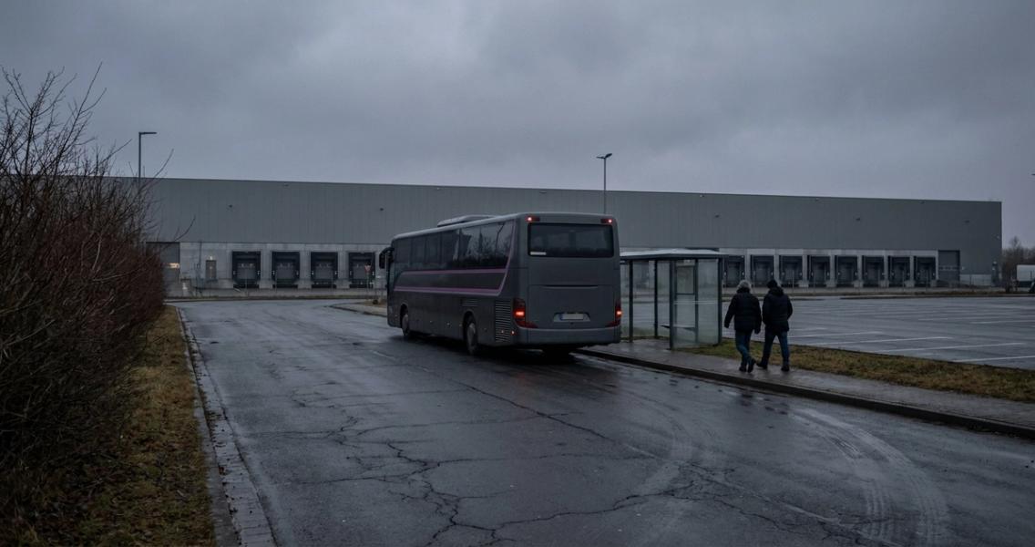 Ein Reisebus wartet an einer leeren Haltestelle vor einem Logistikzentrum, nur zwei Beschäftigte gehen darauf zu