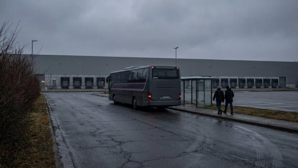Ein Reisebus wartet an einer leeren Haltestelle vor einem Logistikzentrum, nur zwei Beschäftigte gehen darauf zu