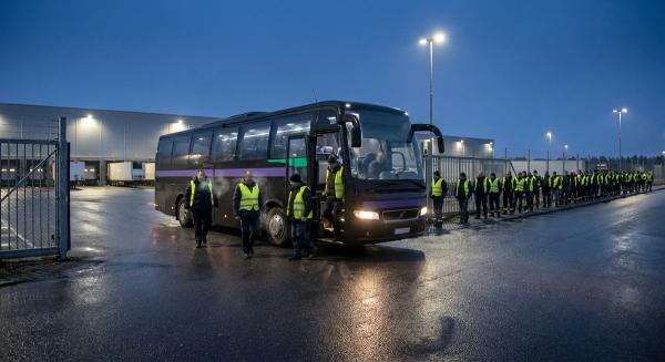 Saisonkräfte steigen vor Sonnenaufgang aus einem Shuttle am Tor eines Versandzentrums, dahinter eine lange Warteschlange