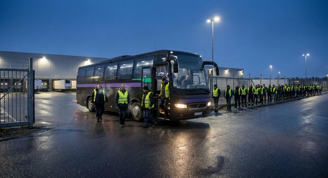Saisonkräfte steigen vor Sonnenaufgang aus einem Shuttle am Tor eines Versandzentrums, dahinter eine lange Warteschlange