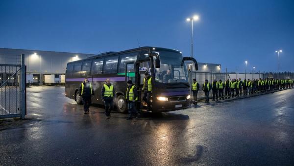 Saisonkräfte steigen vor Sonnenaufgang aus einem Shuttle am Tor eines Versandzentrums, dahinter eine lange Warteschlange