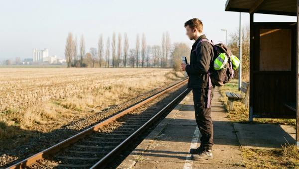 Junger Auszubildender mit Rucksack wartet an einem ländlichen Bahn-Haltepunkt, dahinter Felder und eine Werkshalle