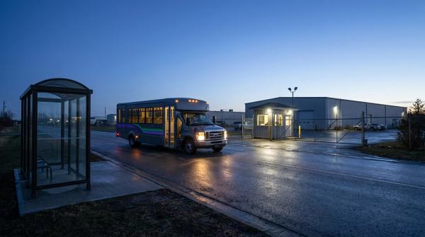 Firmen-Shuttle am Tor eines Gewerbegebiets im Morgengrauen, daneben eine leere, unbeleuchtete Bushaltestelle ohne Linienbus