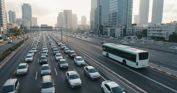 תור רכבים סולו בכביש איילון בשעת שיא בוקר על רקע מגדלי תל אביב, לצד שאטל ארגוני מאוכלס נכנס לפארק הרצליה