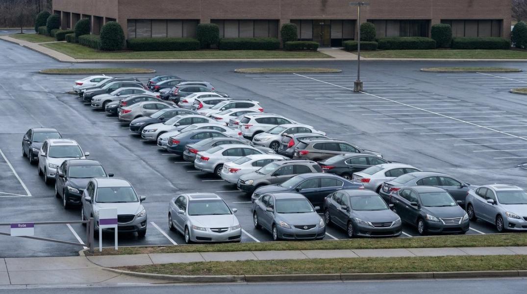 Suburban corporate parking lot full at midday Tuesday with a queue of cars at the entrance, contrasted with empty stalls visible at the back