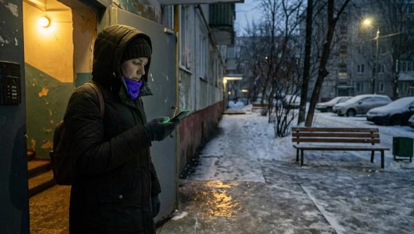 Жінка в зимовій куртці з телефоном у руці стоїть біля дверей під'їзду затемна; поруч обледенілий тротуар