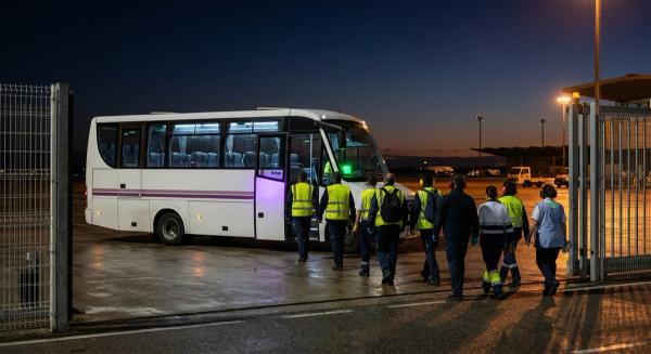 Personal de handling y catering baja de una lanzadera de empresa en el acceso de trabajadores del aeropuerto de madrugada.
