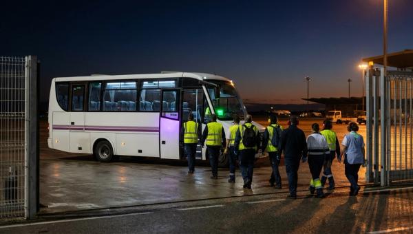 Personal de handling y catering baja de una lanzadera de empresa en el acceso de trabajadores del aeropuerto de madrugada.