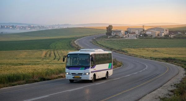 מיניבוס הסעה ארגוני נוסע בכביש כפרי לעבר אזור תעשייה בפריפריה בשעת בוקר מוקדמת, גבעות הגליל ברקע