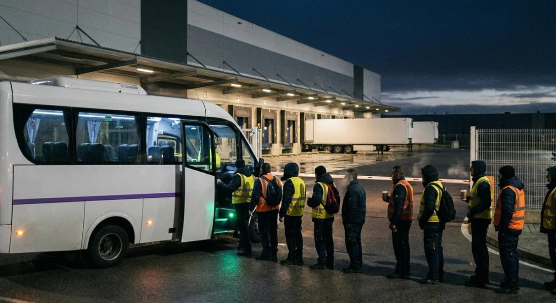 Trabajadores con chaleco reflectante suben a una lanzadera de empresa ante una nave logística de polígono antes del amanecer