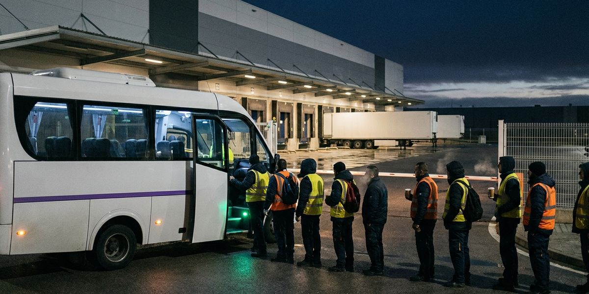 Trabajadores con chaleco reflectante suben a una lanzadera de empresa ante una nave logística de polígono antes del amanecer