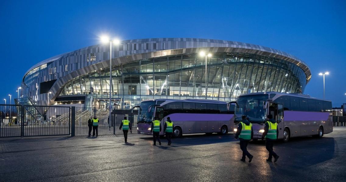 Two staff shuttle buses parked outside an illuminated stadium at dusk as event-day staff in hi-vis vests walk toward them.