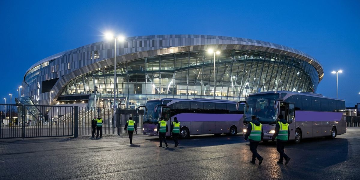 Two staff shuttle buses parked outside an illuminated stadium at dusk as event-day staff in hi-vis vests walk toward them.