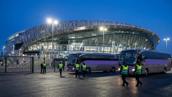Two staff shuttle buses parked outside an illuminated stadium at dusk as event-day staff in hi-vis vests walk toward them.