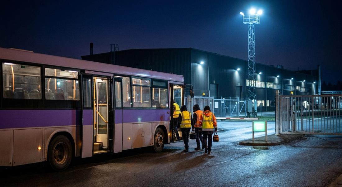 An employee shuttle at a lit manufacturing-plant gate at night as two workers in hi-vis vests board for a shift change