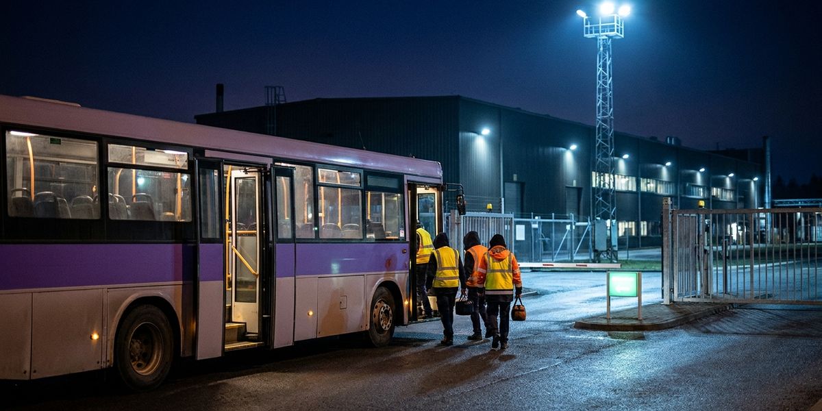 An employee shuttle at a lit manufacturing-plant gate at night as two workers in hi-vis vests board for a shift change