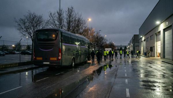 Trabajadores con chalecos reflectantes caminan desde un autocar aparcado hacia una nave industrial al amanecer